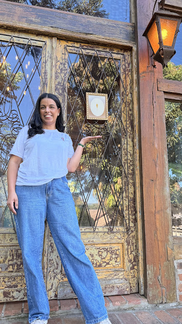 Luana, criadora do @maecomvidaa, sorrindo em frente a uma porta rústica com detalhes em ferro e madeira.